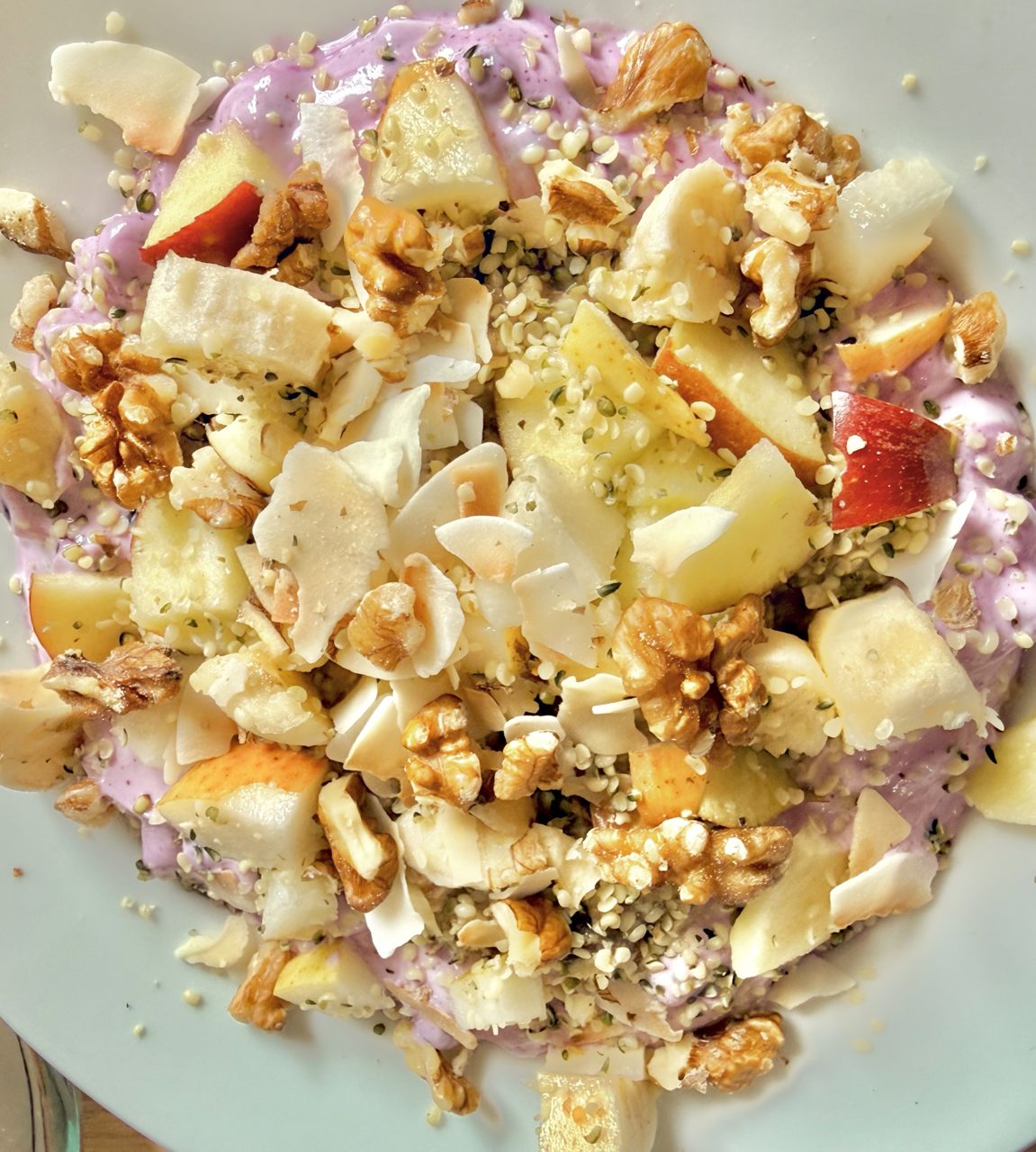 Blue Coconut Bowl: zart lila Heidelbeercreme unter Apfel-, Birnen- und Bananenstücken, mit Kokoschips, Walnuss-Bruch und Hanfsamen in heller Schale