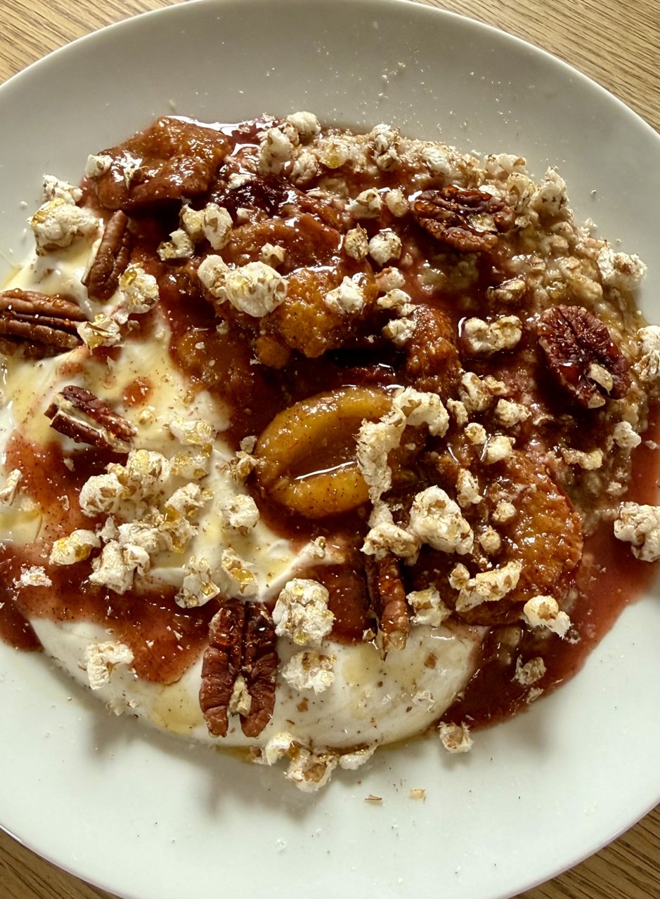 Plum Pecan Bowl: geschmorte Zwetschgen im rötlichen Zimt-Sud auf heller Proteincreme und warmem Porridge, mit Pekannuss-Hälften, Dinkel-Pops und Honigfaden auf weißem Teller