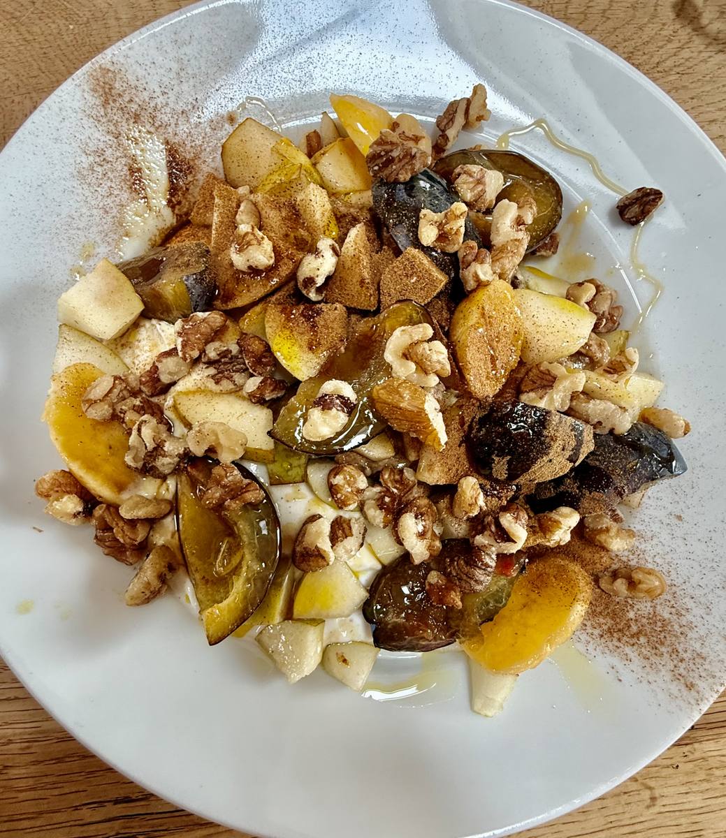 Plummy-Honey Bowl: Zwetschgenspalten, Birne und Mini-Banane auf heller Proteincreme, mit Walnüssen, Hanfsamen, Honigfaden und Ceylon-Zimt auf weißem Teller