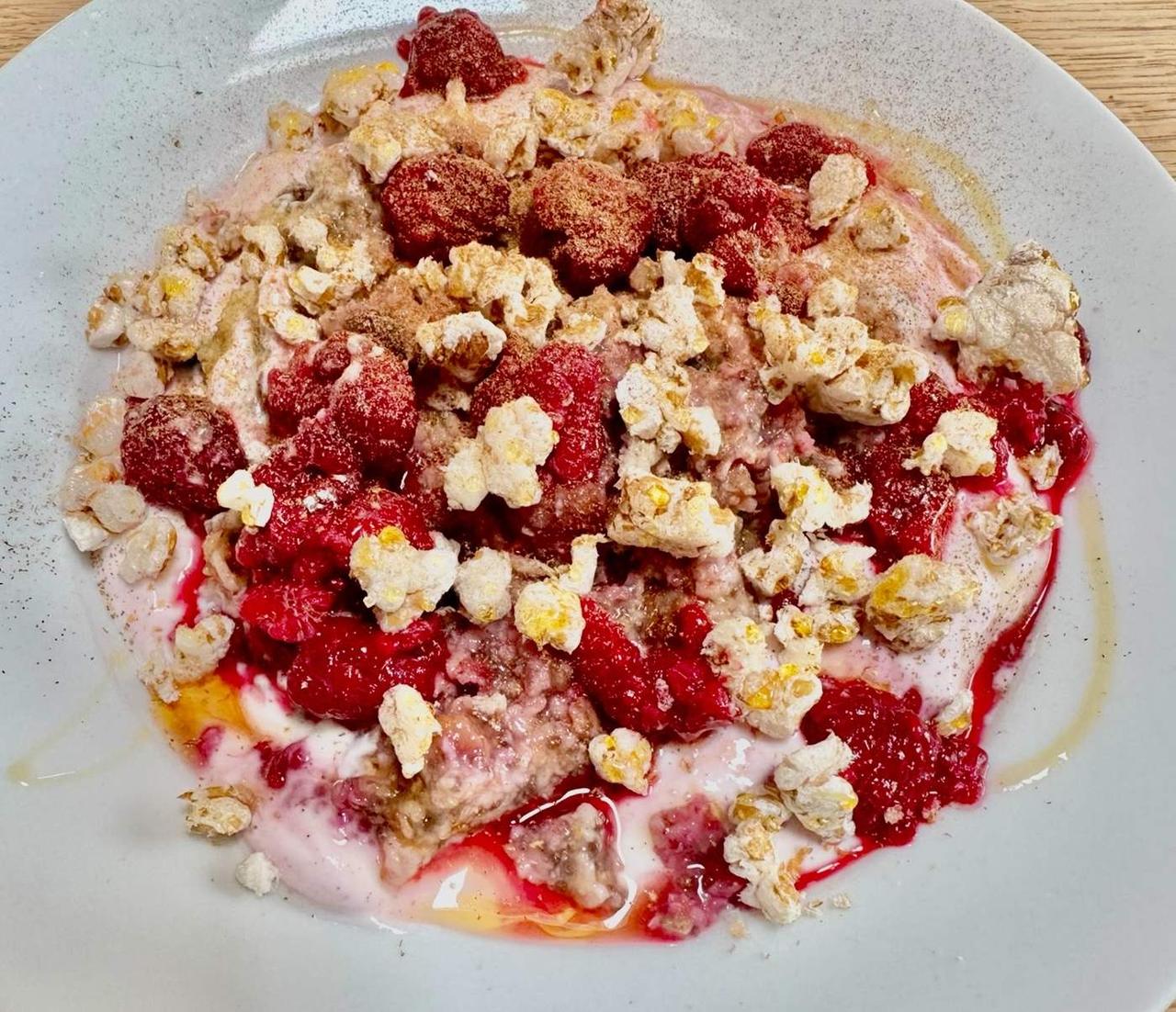 Raspberry Pop Bowl: aufgetaute TK-Himbeeren samt rotem Saft auf rosé Proteincreme, mit goldenen Dinkel-Pops, Honigfaden und Ceylon-Zimt auf grauem Teller