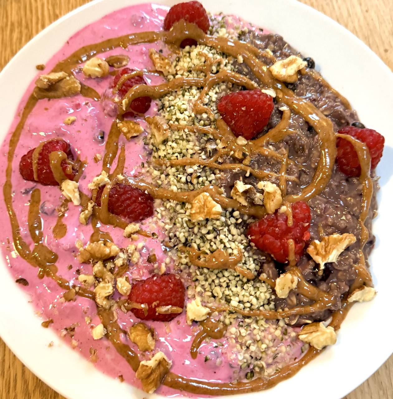 Twin Berry Bowl: links rosé Himbeer-Proteincreme, rechts dunkles Heidelbeer-Feld, getrennt von einem geraden Hanfsamen-Streifen, mit frischen Himbeeren, Walnuss-Bruch und Amlou-Fäden auf hellem Teller